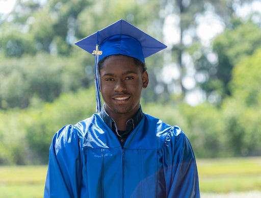 a young black man wearing a blue graduation cap and gown smiles into the camera