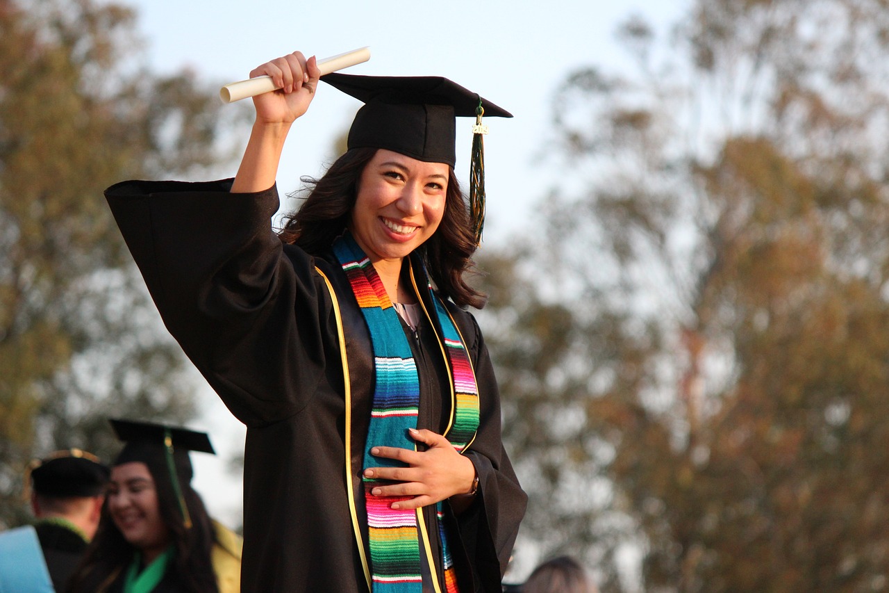 a person wears a black graduation cap and gown. She has a colorful stole and has a diploma in her raised hand, in celebration.